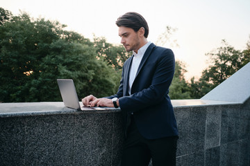 Young attractive brunette man in classic suit standing on street and thoughtfully working on laptop