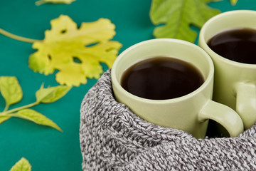 Knitted scarf with two warm  cups of tea, autumn leaves on green background, Autumn. Fall season. Still life.