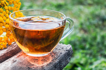 Herbal tea with tansy in a glass mug and yellow tansy flowers on the surface of a wooden table on a background of nature. Tansy Herbal tea. Healing herbs.
