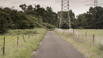 A cyclist passes a high voltage pylon on a road in the meadows.