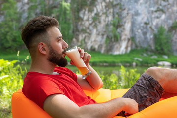 Fototapeta premium A young man with a beard lies on an orange inflatable sofa Lamzak and drinks beer from a glass on the shore. at the picnic enjoys relaxation