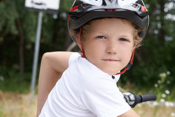 Small Caucasian boy in protective helmet stands leaning on the bike posing for the camera. Teenager ready to ride bicycle in park on summer day. Weekend activity.