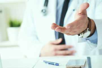Male doctor making welcome gesture, politely inviting patient to sit down in medical office. Photo with depth of field.