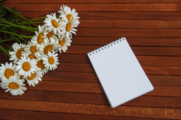 Obraz premium a bouquet of white daisies on a brown wooden table with a clean white notepad, copy space, flat lay, top view.