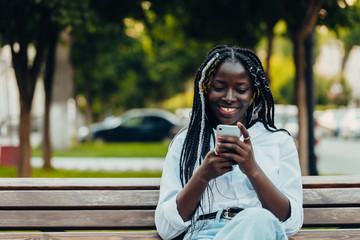 Portrait of a smiling young African American girl with pigtails with a phone sitting on a bench outside on a sunny day. Outdoor photo.