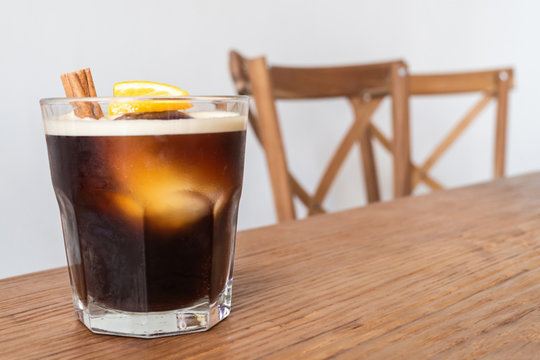 nitro cold brew coffee with ice, cinnamon and lemon on wooden table and chairs set as a special drink menu ready to serve for the client on the coffee shop. cold drink coffee and business concept.