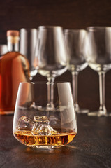 Crystal glass of whiskey with ice on a wooden table. In the background is a full bottle of whiskey and large empty wine glasses.