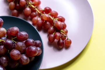 Bunches of ripe purple grapes on the pink plate and in a bowl on a yellow background in the sun. 