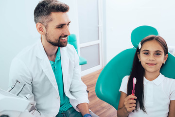 Obraz premium Little mixed race girl in dental chair sitting near her dentist. She holding toothbrush and smiling. The Right Way to Brush Teeth