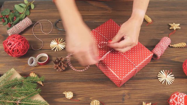 Woman wrapping gift box with equipment and decorating items on wood table, preparing for celebrating Christmas holidays.