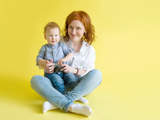 Young beautiful mother with red hair sits in a ginger little son on a yellow background in blue jeans and a white shirt