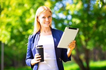 Fototapeta premium Young happy smiling caucasian adult girl business student walking concept in the park holding reading tablet thinking of credit finance job in the college with wireless headphones drink coffee