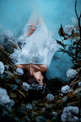 A woman lies on the ground in a short dress with bare shoulders and she is gently covered by a cold, dense fog, like a blanket. Background blooming hydrangea and haze. Art processing of photography