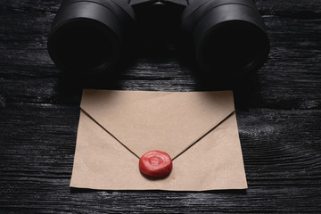 Binoculars and a blank envelope with a sealing wax on a black wooden board background.