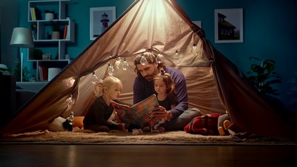 Happy dad and little daughters read a book in a tent at home