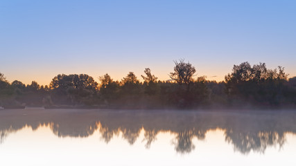 Panorama of tendrils of morning mist on a lake at sunrise and a colorful orange glow in the sky reflected in the tranquil water with the surrounding woodland trees in an atmospheric landscape
