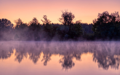 Panorama of tendrils of morning mist on a lake at sunrise and a colorful orange glow in the sky reflected in the tranquil water with the surrounding woodland trees in an atmospheric landscape