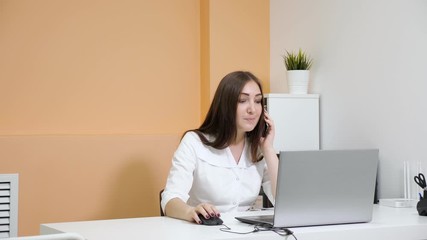 smiling lady receptionist in white coat talks on smartphone explaining to customer price list at laptop in beauty salon