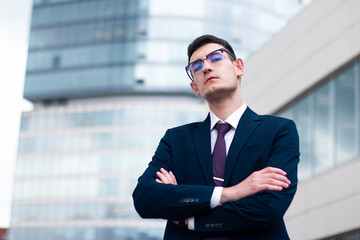 Serious arrogant young businessman in a suit with tie and in glasses looking down at camera and holding his hands or arms crossed standing outdoor in front of modern office business centre, skyscraper