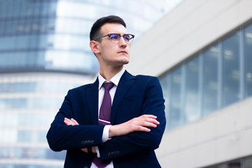 Confident serious young businessman in a formal suit and in glasses liking into distance and holding his hands or arms crossed, standing outdoor near modern office centre. Business people concept.