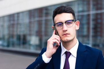 Handsome young businessman in glasses talking on cell phone and looking at camera, wearing suit. Serious business entrepreneur in jacket, shirt and tie having conversation on smartphone outdoors.