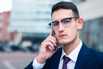 Serious confident young businessman in glasses and suit looking at camera close up and talking on cell phone. Smart dressed guy having call, conversation on smartphone, in negotiations outdoor office