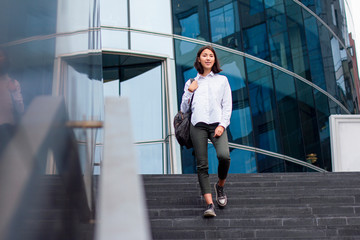Young asian businesswoman or student goes down the stairs with a backpack from the work place or office. Formal dressed girl in white shirt with braces smiling and ending her work job day, outdoor