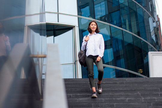 Young asian businesswoman or student goes down the stairs with a backpack from the work place or office. Formal dressed girl in white shirt with braces smiling and ending her work job day, outdoor - Powered by Adobe