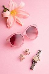 Glamorous woman accessories - watch, sunglasses, earrings and lily flower. Feminine composition on pink background