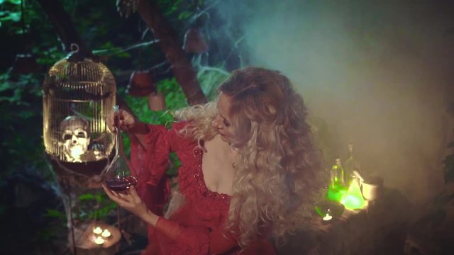 Mysterious blond woman in a red baroque dress with bare shoulders and lace. She holds a jar of red liquid in her hands and shakes it. Background of a night witch's yard with smoke and fog.