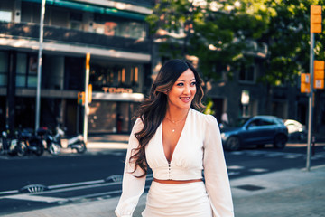 Fototapeta premium Summer sunny lifestyle portrait of young stylish asian girl on the street of big modern city. She wears white dress