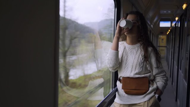 Young girl travelling on train in daytime.Wide angle view charming woman standing in passage and enjoying landscape view passing by out of window. She brooding drinks coffee near the big window 