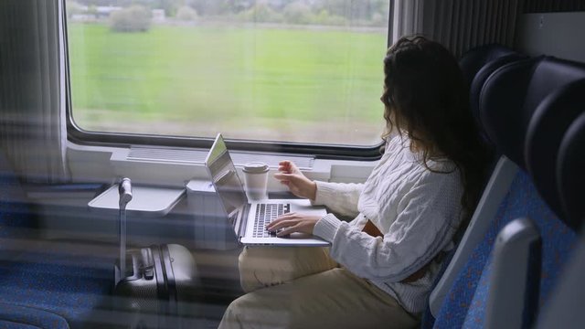 Young girl travelling on train in daytime. Side view charming woman sitting near window, working on computer. Female drinking coffee and enjoying landscape view passing by at background. 