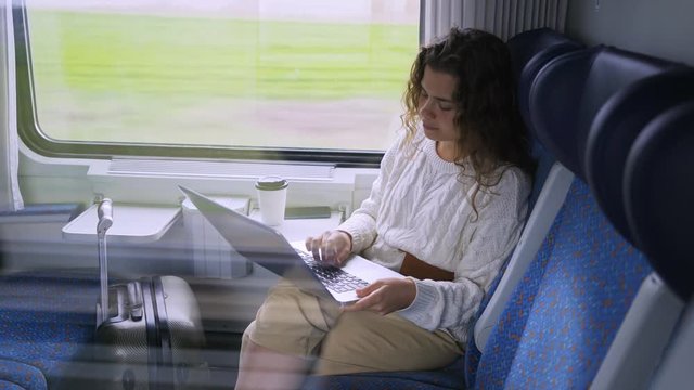Young girl travelling on train in daytime. Charming woman sitting near window, working on computer. Female drinking coffee typing on laptop and enjoying landscape view passing by at background. 