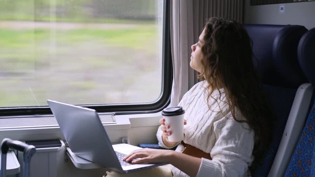 Young girl travelling on train in daytime. Crop view woman sitting near window, working on computer. Female drinking coffee typing on laptop and enjoying landscape view passing by at background. 