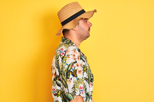 Young man on vacation wearing summer shirt and hat over isolated yellow background looking to side, relax profile pose with natural face with confident smile.