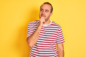 Young man wearing casual striped t-shirt standing over isolated yellow background with hand on chin thinking about question, pensive expression. Smiling with thoughtful face. Doubt concept.