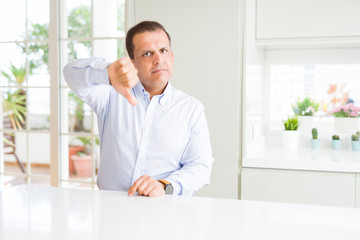 Middle age man sitting at home looking unhappy and angry showing rejection and negative with thumbs down gesture. Bad expression.