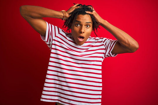 Young afro man with dreadlocks wearing striped t-shirt standing over isolated red background Crazy and scared with hands on head, afraid and surprised of shock with open mouth