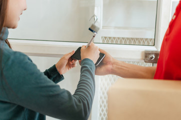 young woman customer appending digital signature in mobile phone receiving parcel post box from courier with home delivery service man smiling face in red uniform at home, express delivery concept
