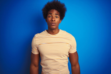 Fototapeta na wymiar American man with afro hair wearing striped yellow t-shirt over isolated blue background with serious expression on face. Simple and natural looking at the camera.