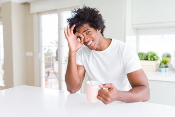 African American man with afro hair drinking a cup of coffee with happy face smiling doing ok sign with hand on eye looking through fingers