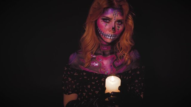 bright lady with color makeup in style of day of dead, woman with rhinestones on face in image of St. Katrina, sweet pink and violet shiny cheper with burning candle in hands with open shoulders