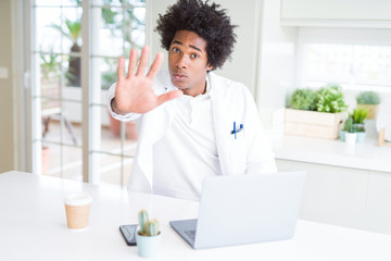 African American doctor man working with laptop at the clinic with open hand doing stop sign with serious and confident expression, defense gesture