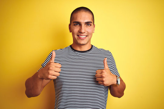 Young handsome man wearing striped t-shirt over yellow isolated background success sign doing positive gesture with hand, thumbs up smiling and happy. Cheerful expression and winner gesture.