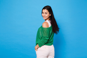 Back rear view of attractive young woman in green casual clothes looking back, holding hands on pockets isolated on bright blue wall background in studio. People lifestyle concept. Mock up copy space.