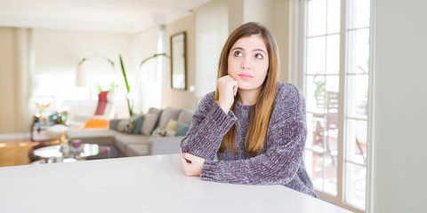 Beautiful young woman at home with hand on chin thinking about question, pensive expression. Smiling with thoughtful face. Doubt concept.