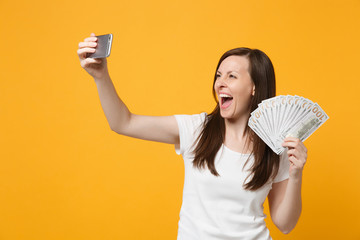 Joyful young woman in white clothes doing selfie shot on mobile phone, holding fan of cash money in dollar banknotes isolated on yellow orange background. People lifestyle concept. Mock up copy space.