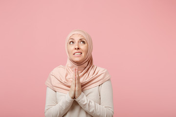 Pensive young arabian muslim woman in hijab light clothes posing isolated on pink background in studio. People religious Islam lifestyle concept. Mock up copy space. Holding hands folded in prayer.