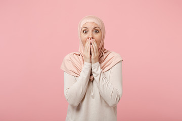 Shocked young arabian muslim woman in hijab light clothes posing isolated on pink background studio portrait. People religious Islam lifestyle concept. Mock up copy space. Covering mouth with hands.